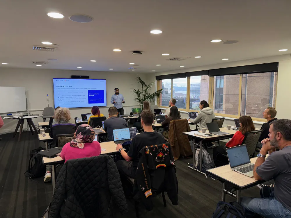 Product people at laptops in a Sydney meeting room, the Opera House through the windows, as Kailash introduces Claude Code on the screen